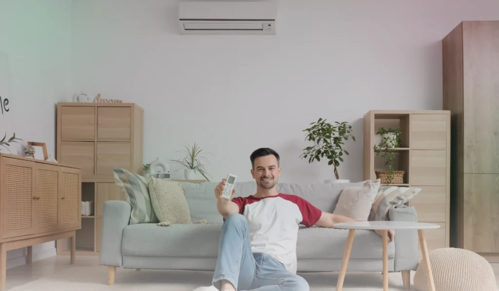 Man smiling at the camera holding an air conditioner remote control with the AC unit mounted on the wall.