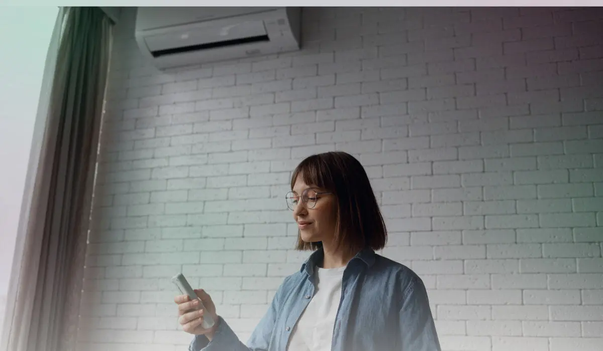 Happy woman relaxing on a couch holding an AC remote, with an air conditioner mounted on the wall.