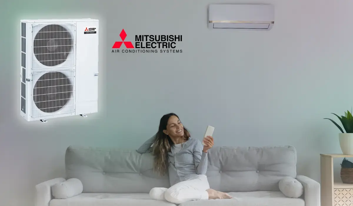 Happy woman relaxing on a couch holding an AC remote, with a Mitsubishi air conditioner mounted on the wall.